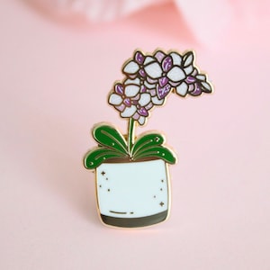 May include: Gold enamel pin featuring a white orchid with purple accents in a white pot with gold accents and a speckled design.