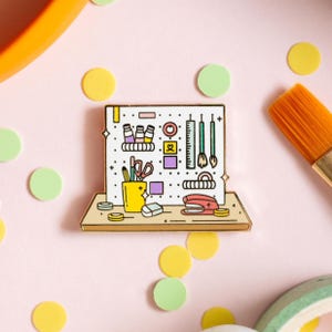 May include: A gold-toned enamel pin depicting a white pegboard with various office supplies, including pencils, scissors, paintbrushes, a stapler, tape, and a ruler. The pegboard is mounted on a brown wooden shelf.