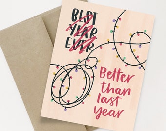 May include: A pink card with a black and white illustration of string lights. The text on the card reads "Best year ever" with "ever" crossed out and "Better than last year" written below.