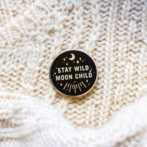 K&ouml;nnte beinhalten: Schwarzer Emaille-Pin mit goldener Aufschrift "Stay Wild Moon Child". Der Pin zeigt einen Halbmond und Sterne.