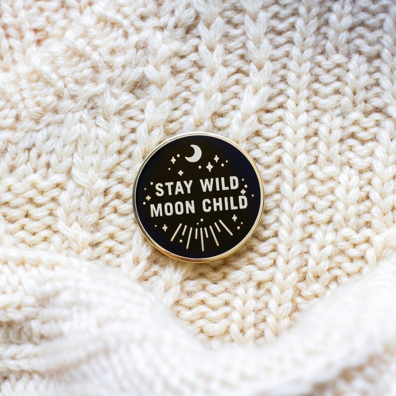 K&ouml;nnte beinhalten: Schwarzer Emaille-Pin mit goldener Aufschrift "Stay Wild Moon Child". Der Pin zeigt einen Halbmond und Sterne.