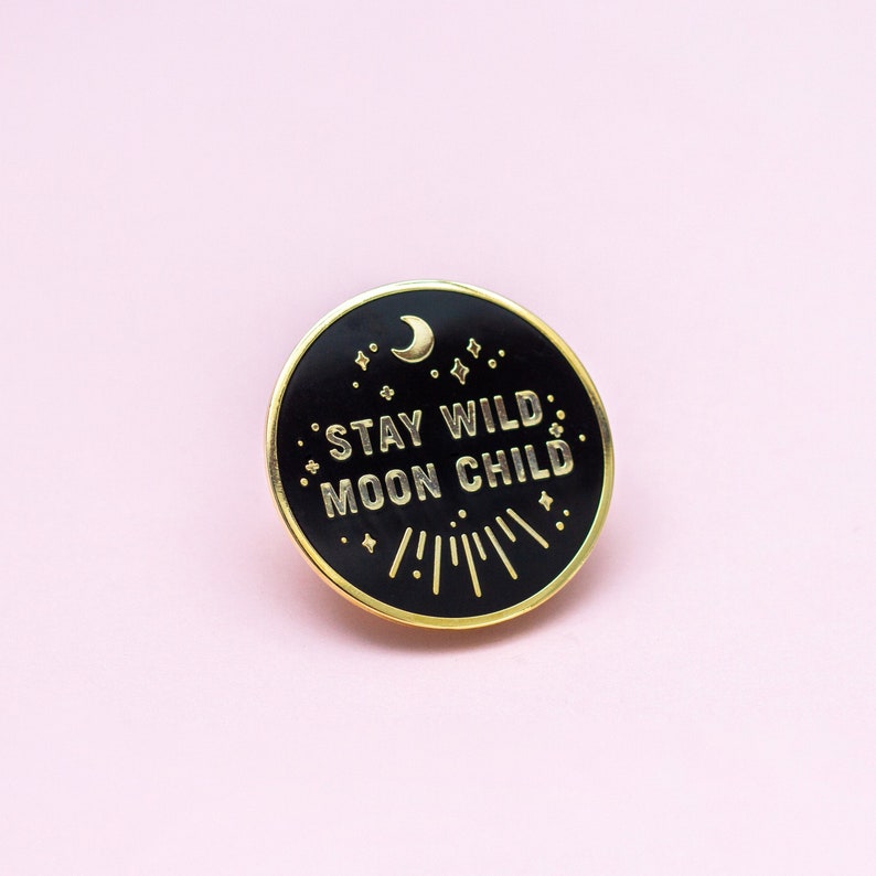 K&ouml;nnte beinhalten: Schwarzer Emaille-Pin mit goldener Aufschrift "Stay Wild Moon Child". Der Pin zeigt einen Halbmond und Sterne.