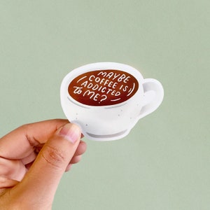 May include: White sticker in the shape of a coffee cup, filled with brown liquid. The text "MAYBE COFFEE IS ADDICTED TO ME?" is written in white. The sticker is held by a hand against a light green background.