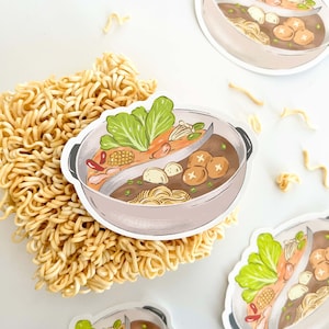 May include: A sticker of a hot pot with a variety of ingredients, including vegetables, meat, and noodles. The hot pot is divided into two sections, one with a clear broth and the other with a darker broth. The sticker is on top of a pile of instant noodles.