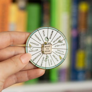 Puede incluir: Un pin de color dorado con forma de rueda con un icono de libro en el centro que dice "What to Read". La rueda tiene 12 secciones con diferentes géneros de libros listados, incluyendo fantasía, horror, thriller, ciencia ficción, memorias, romance, misterio, novela gráfica, poesía, ficción, ficción histórica y autoayuda.