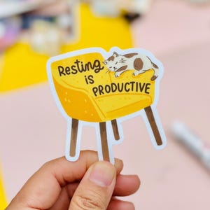 May include: A sticker featuring a yellow cartoon chair with brown legs and the text "Resting is Productive." A brown and white cat is sleeping on the chair. The sticker is held by a hand.