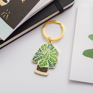 May include: Gold-tone keychain featuring a green and white enamel plant with a brown pot. The plant has a leafy design and is attached to a gold-tone key ring.