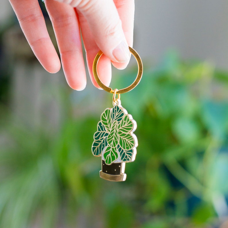 May include: Gold keychain featuring a green and white enamel plant with a gold pot. The plant has a rounded shape and resembles a leafy houseplant.