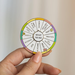 May include: A gold-coloured metal pin shaped like a wheel with a white centre. The wheel is divided into 12 sections, each with a different activity written on it. The activities include "Call a Friend", "Clean", "Exercise", "Eat", "Watch TV", "Read", "Go Outside", "Meditate", "Nap", "Arts and Crafts", "Shower", and "Play a Game". The centre of the wheel says "What to Do".