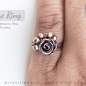 Puede incluir: Un anillo de alambre de cobre envuelto con un diseño de rosa. El anillo está en un dedo. La rosa está hecha de alambre de cobre y tiene un centro negro. El anillo está rodeado de pequeñas cuentas de cobre.