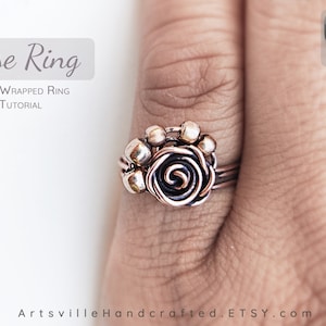 Pu&ograve; includere: Un anello avvolto in filo di rame con un design a forma di rosa. L'anello &egrave; indossato su un dito. La rosa &egrave; realizzata in filo di rame e ha un centro nero. L'anello &egrave; circondato da piccole perline di rame.