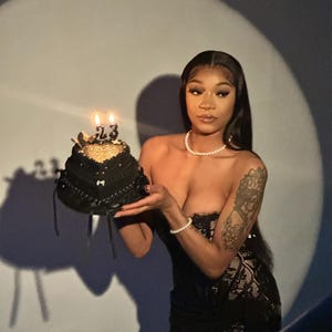 Op de afbeelding: Een zwarte verjaardagstaart met gouden accenten en twee brandende kaarsen met het nummer 23. De taart wordt vastgehouden door een persoon die een zwarte strapless jurk en een parelketting en armband draagt.