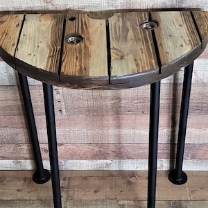 May include: A half-circle wooden table with a rustic finish and black metal legs. The table top is made from reclaimed wood and has a natural wood grain pattern.