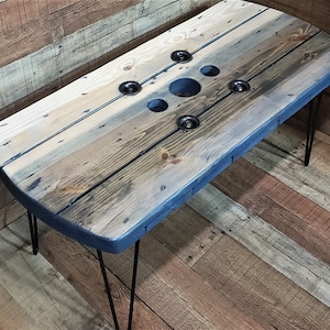 May include: A rustic coffee table made from a repurposed wooden cable spool. The tabletop features a mix of light and dark wood tones, with metal accents and black hairpin legs. The table has a blue painted edge and several circular holes.