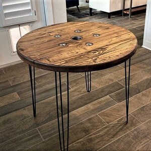 May include: A round wooden table with a rustic finish and a hole in the center. The table is supported by four black hairpin legs.