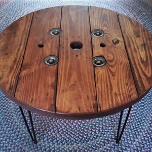 May include: A round wooden table with a dark brown stain. The table top is made from a repurposed cable spool and has a natural wood grain pattern. The table has hairpin legs made of black metal.