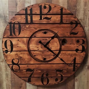 May include: A wooden clock with black Roman numeral markings on a brown wooden background. The clock face is made of wood and has a black clock hand.