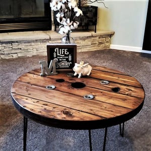 May include: A round wooden coffee table made from a repurposed cable spool with black hairpin legs. The table top has a dark brown wood stain and features several holes. A small white pig figurine and a sign that reads "Life is better in the country" are on the table.