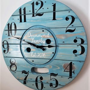May include: A blue wooden clock with black numbers and hands. The clock is made from a repurposed wooden spool and has a white inscription that reads "Drummond Family Est. 2016".