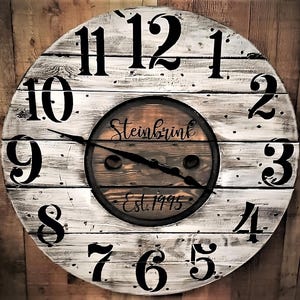 May include: A large, whitewashed wooden clock with black Roman numeral hour markers. The clock face is made from reclaimed wood and has a brown wooden center with the text "Steinbrink Est. 1995".