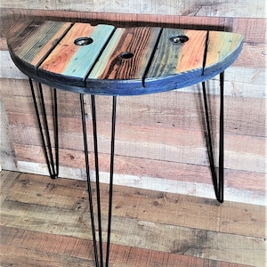 May include: A half-round wooden table with a distressed, multi-colored finish. The table has a black stain and is supported by four hairpin legs.