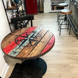 May include: A round wooden table with a compass design painted on the top. The table has a black metal base and is surrounded by a light wood floor.