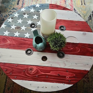 May include: A round wooden coffee table painted with the American flag design. The table features red and white stripes, a blue section with white stars, and decorative items including a candle, a green ceramic bird, and a small green plant.