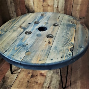 May include: A round wooden coffee table made from a repurposed cable spool. The table has a dark blue stain and is supported by three hairpin legs.