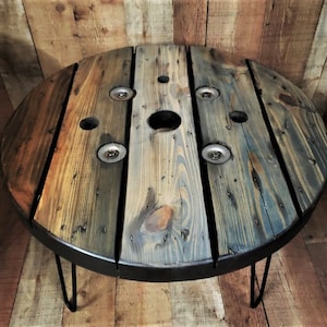 May include: A round wooden coffee table with a black metal base and hairpin legs. The table top is made from reclaimed wood and has a distressed finish. The table top has several holes and metal accents.