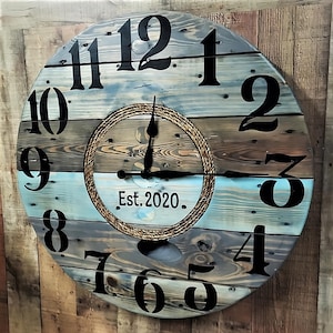 May include: A large, round, wooden clock with a rope design around the center. The clock face is painted in shades of blue, brown, and white, with black numbers and hands. The clock reads "Est. 2020."