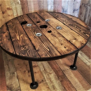 May include: A round wooden table with a dark brown stain. The table top is made from a cable spool and has a rustic look. The table has three black metal legs.