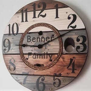 May include: A rustic wooden clock with Roman numeral hour markers and a rope design around the center. The clock face is made of reclaimed wood planks in shades of brown, gray, and white. The clock face also includes the text "Benner Family".