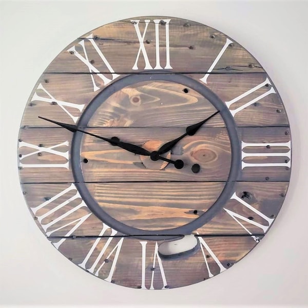 Könnte beinhalten: Eine große, runde Holzuhr mit römischen Ziffern in weißer Farbe. Das Zifferblatt der Uhr ist aus wiederverwendetem Holz und hat einen grau lackierten Ring am Rand. Die Zeiger der Uhr sind schwarz.