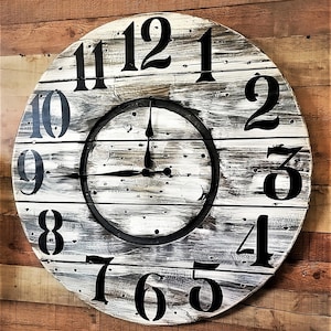 May include: A large, round wall clock with a distressed, whitewashed wooden face. Bold black numerals mark the hours. The clock hands are black, and the clock is mounted on a wooden wall. The clock's diameter is approximately 24 inches.