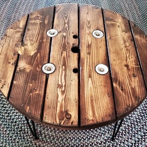 May include: A round wooden table made from a repurposed cable spool. The table has a dark brown stain and is supported by hairpin legs. The table top has a natural wood grain pattern and features four metal accents.