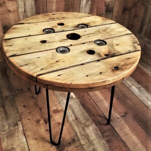 May include: A round wooden table with a natural wood finish. The table top is made from a repurposed cable spool and has several holes. The table is supported by three black hairpin legs.
