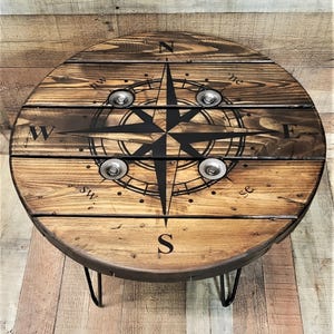 May include: A round wooden table featuring a dark brown stained finish with a black compass rose graphic printed on the surface. The design includes the cardinal directions N, E, S, W, and intermediate points, with four silver metal bolts.