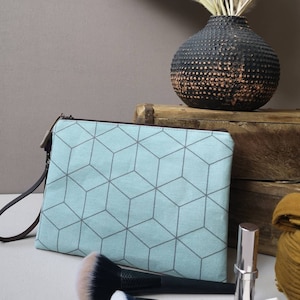 Könnte beinhalten: Eine hellblaue Kosmetiktasche aus Canvas mit geometrischem Muster und einem braunen Lederriemen. Die Tasche liegt auf einer Holzoberfläche mit einer Vase mit getrockneten Blumen im Hintergrund.