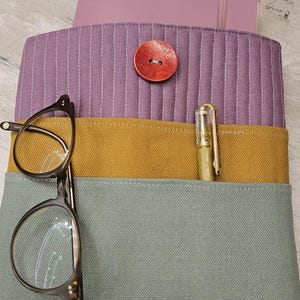 Peut inclure: Un organisateur fait à la main en nuances de violet, jaune moutarde et sarcelle. L'organisateur comporte une fermeture à bouton rouge, une paire de lunettes et un stylo doré. L'étiquette indique "ERIKA LANG Handmade Carries a Soul."