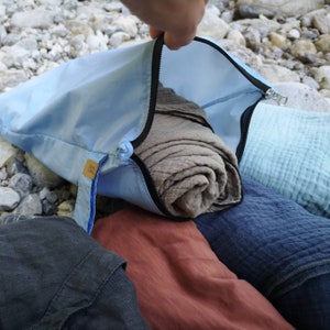 Może przedstawiać: Jasnoniebieska torba zapinana na zamek zawiera zwinięty, beżowy koc. Widoczne są inne złożone koce w odcieniach niebieskiego, czerwonego i szarego. Torba ma małą, prostokątną metkę z tekstem. Scena rozgrywa się na zewnątrz na skalistej powierzchni.