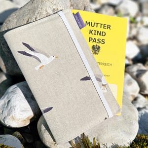 May include: A beige fabric journal cover with a white elastic band and a printed design of seagulls in flight. The cover is placed over a yellow document with the text "MUTTER KIND PASS". The background includes rocks and greenery.