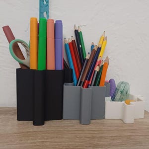 Op de afbeelding: Bureau-organizers gevuld met diverse briefpapierartikelen. De zwarte, grijze en witte organizers bevatten kleurrijke stiften, potloden, scharen en een tape dispenser. De items zijn geplaatst op een houten oppervlak.