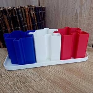 Può includere: Tre contenitori a forma di puzzle, blu, bianco e rosso, sono disposti in fila su una base bianca. I contenitori sono in plastica e hanno una finitura liscia e lucida.