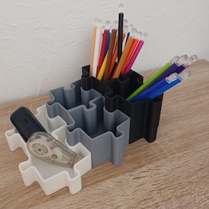 Peut inclure: Un organiseur de bureau à compartiments multiples en forme de pièce de puzzle. L'organisateur est blanc, gris et noir, contenant des crayons et des stylos de différentes couleurs, une télécommande noire et un correcteur. Il est posé sur une surface en bois.