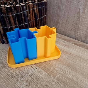 May include: A blue and yellow puzzle-shaped desktop organizer with two compartments. The organizer is made of plastic and has a smooth finish.