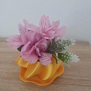 May include: A small, yellow, abstract-shaped vase holding pink flowers with dark markings and green and white accents. The vase sits on a matching yellow base, creating a modern floral arrangement.
