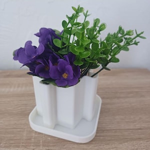 May include: A white, star-shaped flower pot with a matching saucer, filled with vibrant purple artificial flowers and green foliage. The pot sits on a light brown wooden surface, creating a simple, yet elegant display.