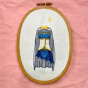 May include: Cross-stitch embroidery of a blue and white fantasy dress with a cape, set in an oval wooden frame. The dress features gold accents and is centered on a white fabric background. The frame is secured with a brass clasp.