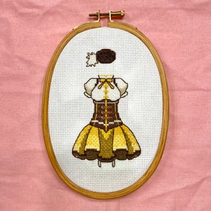 May include: A cross-stitch embroidery of a yellow and brown dress with a white blouse and a white bow. The dress is framed in a wooden hoop.