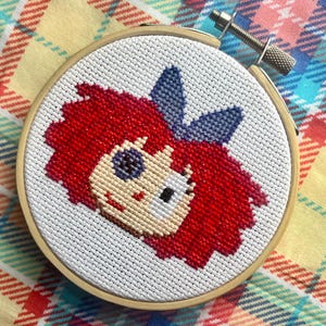 Puede incluir: Un bastidor de bordado de punto de cruz con la cara de una muñeca con pelo rojo y un lazo azul. La cara de la muñeca es beige, con ojos negros y boca blanca. El bordado está en un bastidor de madera, sobre un fondo a cuadros.